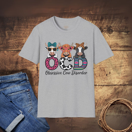 Gray t-shirt with colorful cow design and 'Obsessive Cow Disorder' text on a wooden surface with jeans and rope.