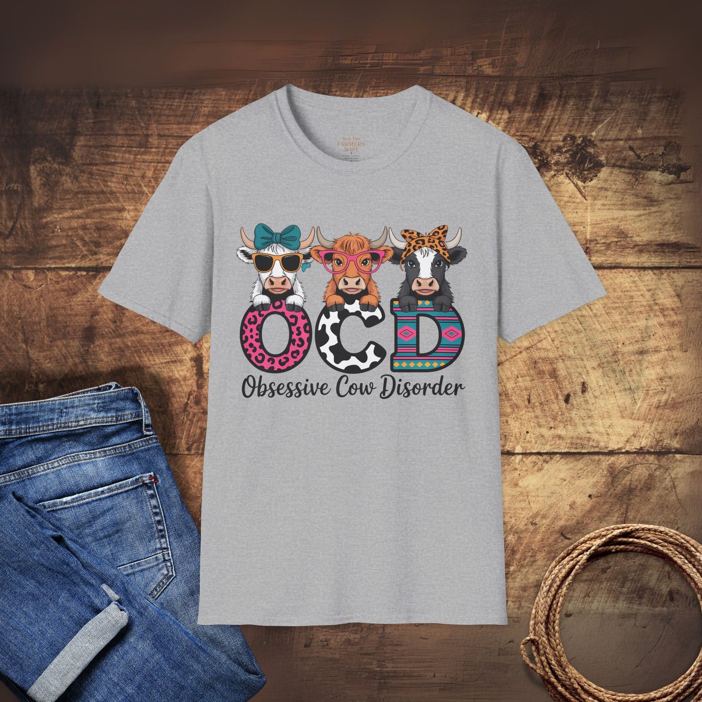 Gray t-shirt with colorful cow design and 'Obsessive Cow Disorder' text on a wooden surface with jeans and rope.