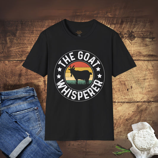 Black t-shirt with 'The Goat Whisperer' design on a wooden surface with jeans and a bowl of goat cheese.